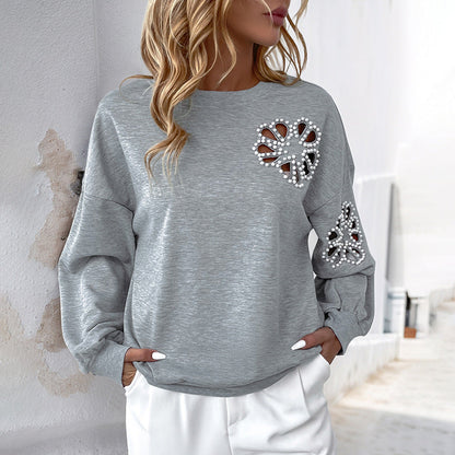 Frau in grauem Pullover mit Perlenherz-Design, lässig und modisch, steht vor weißer Wand. Trendige Damenmode, stilvolles Outfit, Freizeitlook.