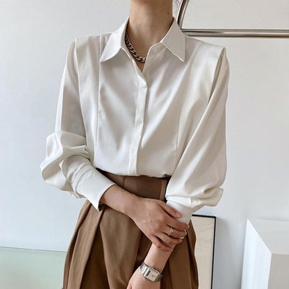 Frau in eleganter weißer Bluse mit langen Ärmeln und braunem Rock, trägt eine silberne Halskette und Armbanduhr. Businessmode, Damenbekleidung.