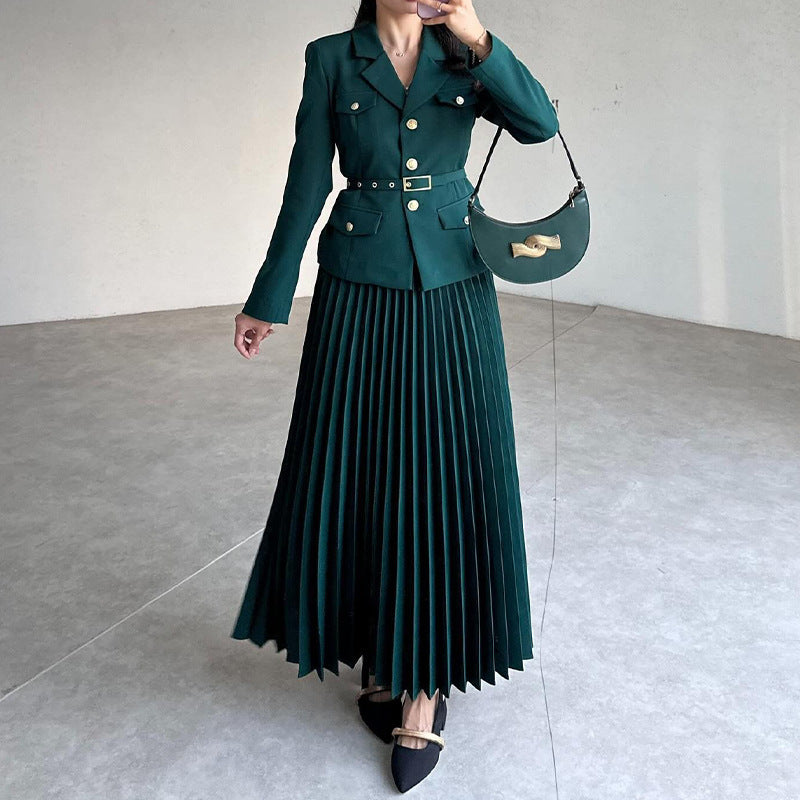 Frau in elegantem, grünem, plissiertem Maxikleid mit Gürtel und goldenen Knöpfen, trägt schwarze spitze Schuhe und eine stilvolle Handtasche.