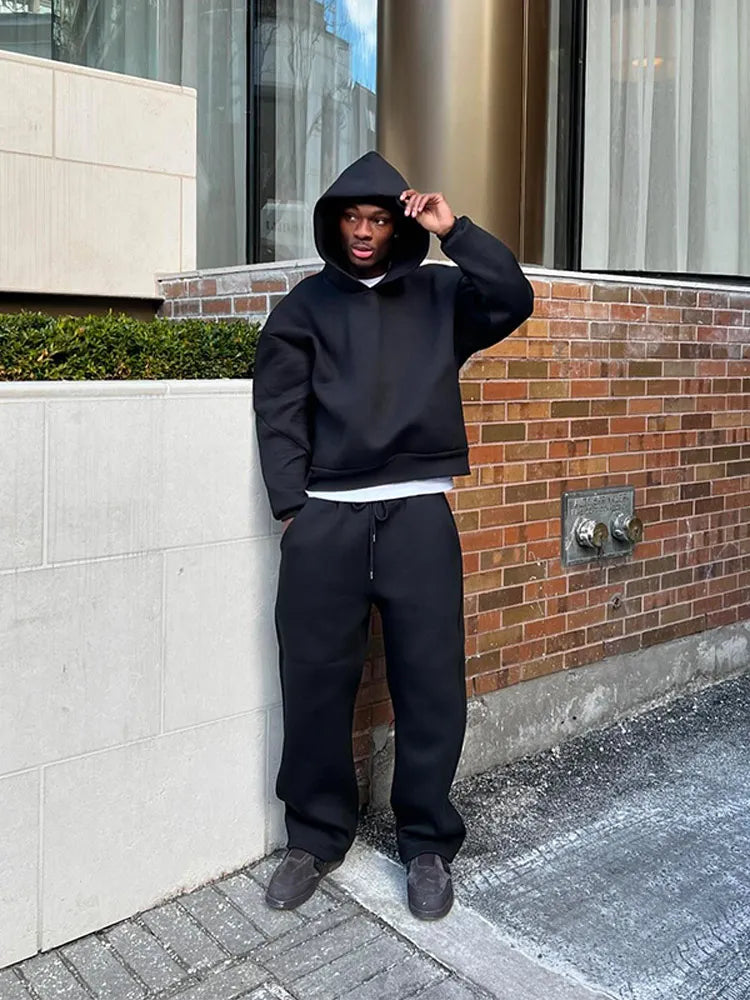 Ein Mann in schwarzem Hoodie und Jogginghose steht vor einer Backsteinmauer. Urbaner Streetwear-Look, lässiger Stil, moderne Mode.