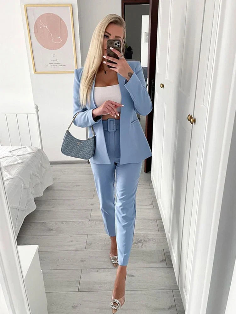 Blonde Frau im blauen Anzug macht Selfie im Spiegel. Eleganter Business-Look mit weißem Top, modischer Handtasche und Absatzschuhen.