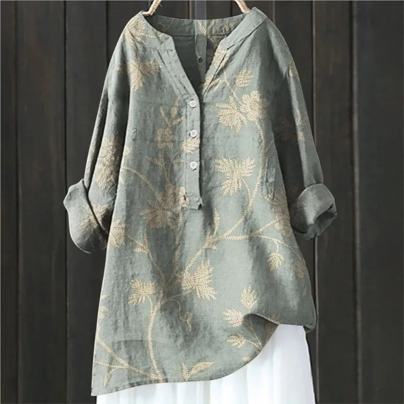 Graues Leinenhemd mit Blumenmuster, langen Ärmeln und Knopfleiste, hängend vor dunklem Holzhintergrund. Elegante Damenmode, lässiger Stil.
