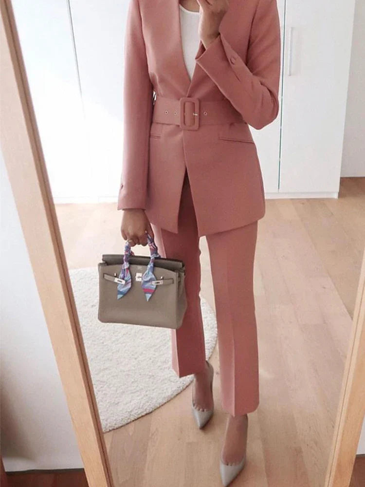 Frau in rosa Anzug mit Gürtel, hält graue Handtasche mit Seidenschal, steht vor Spiegel. Businessmode, eleganter Stil, modische Accessoires.