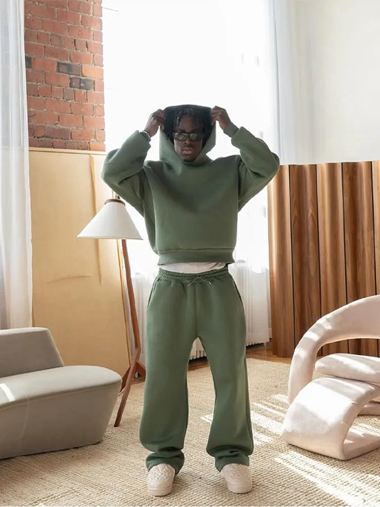 Person in grünem Hoodie und Jogginghose steht in modernem Wohnzimmer mit Stehlampe und Designerstuhl. Lässige Mode, urbaner Stil, gemütliche Einrichtung.