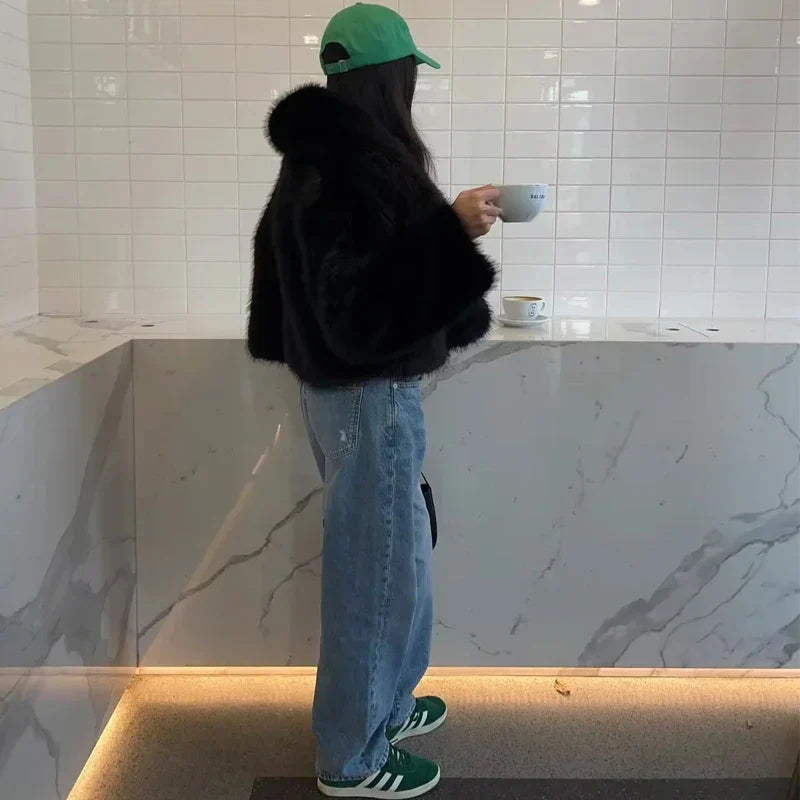 Frau in schwarzer Felljacke und blauer Jeans, trägt grüne Kappe und Sneakers, hält Tasse in modernem Café mit Marmortresen und weißen Fliesen.