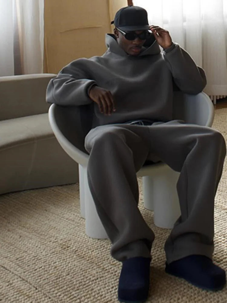Mann in grauem Oversize-Sweatshirt und Jogginghose sitzt auf modernem Stuhl, trägt schwarze Sonnenbrille und Mütze, lässiger Streetwear-Look.