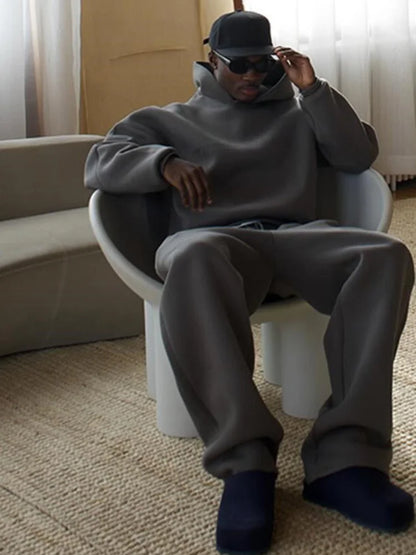 Mann in grauem Oversize-Sweatshirt und Jogginghose sitzt auf modernem Stuhl, trägt schwarze Sonnenbrille und Mütze, lässiger Streetwear-Look.