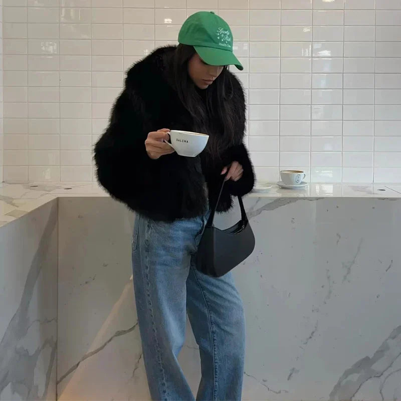 Frau in schwarzem Pelzmantel und grüner Kappe trinkt Kaffee in modernem Café. Marmor-Theke, lässiger Stil, modische Accessoires.