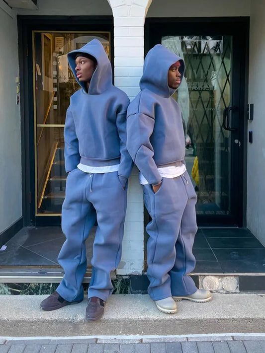 Zwei Personen in blauen Oversize-Hoodies und Jogginghosen stehen Rücken an Rücken vor einem Gebäude. Streetwear, lässiger Stil, urbane Mode.