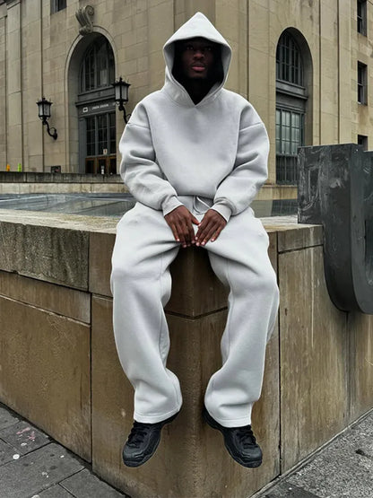 Mann in grauem Hoodie und Jogginghose sitzt auf Mauer vor historischem Gebäude. Streetwear, urbaner Stil, lässige Mode, Stadtlandschaft.