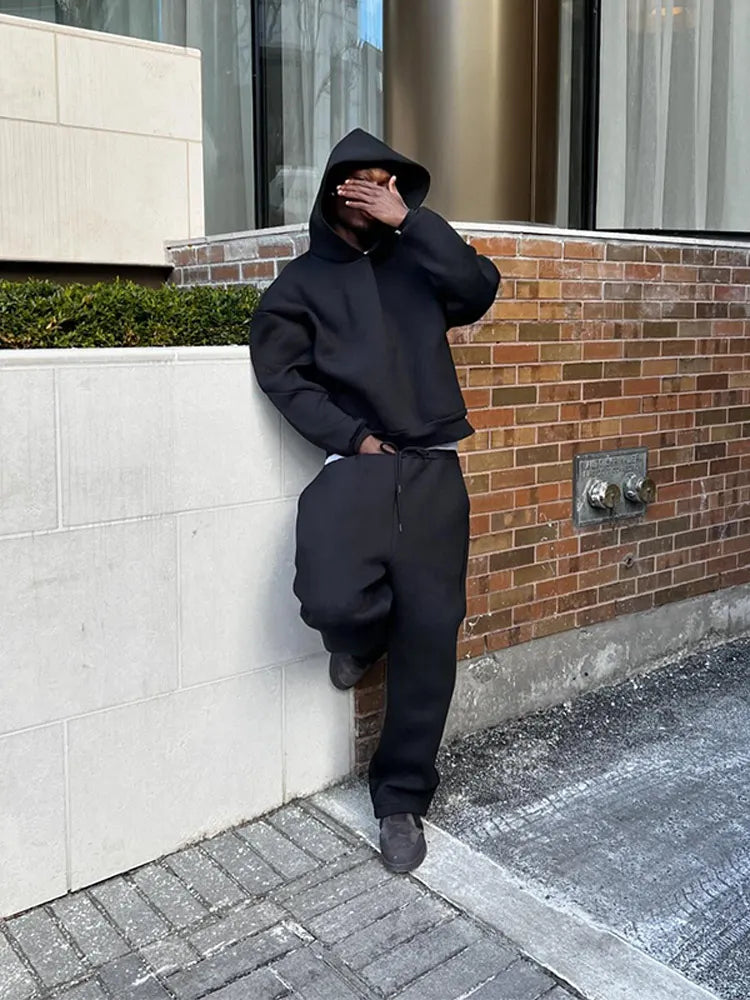 Person in schwarzem Hoodie und Jogginghose lehnt an einer Backsteinmauer, verdeckt Gesicht mit Hand. Urbaner Streetstyle, lässige Mode.