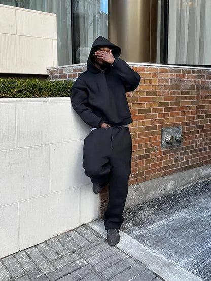 Person in schwarzem Hoodie und Jogginghose lehnt an einer Backsteinmauer, verdeckt Gesicht mit Hand. Urbaner Streetstyle, lässige Mode.