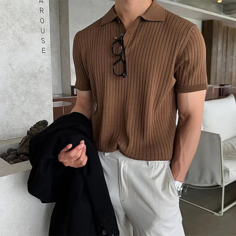 Mann in braunem, geripptem Poloshirt mit Brille am Kragen, hält schwarze Jacke, trägt weiße Hose. Modernes, stilvolles Herrenoutfit.