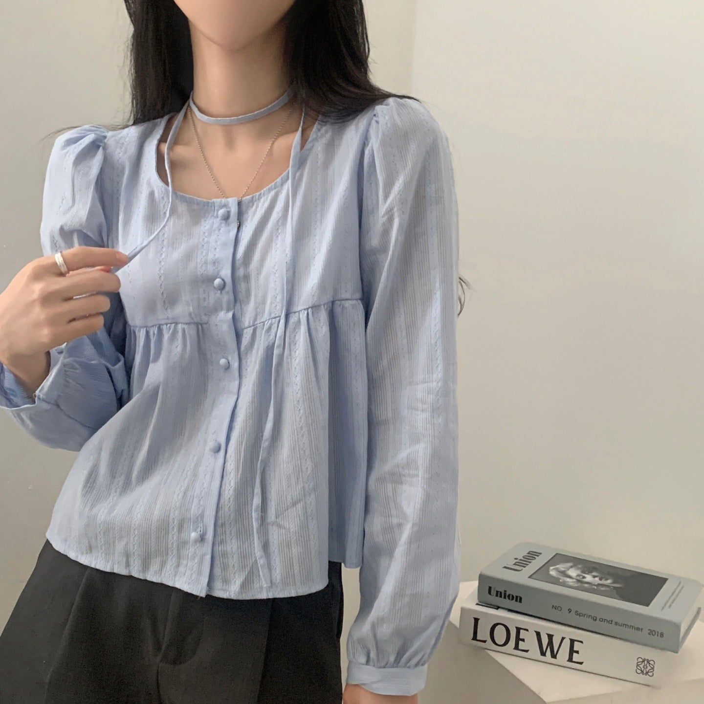 Frau in hellblauer Bluse mit Knopfleiste, hält Kordel. Schwarze Hose, Bücherstapel im Hintergrund. Modetrend, Damenmode, eleganter Look.