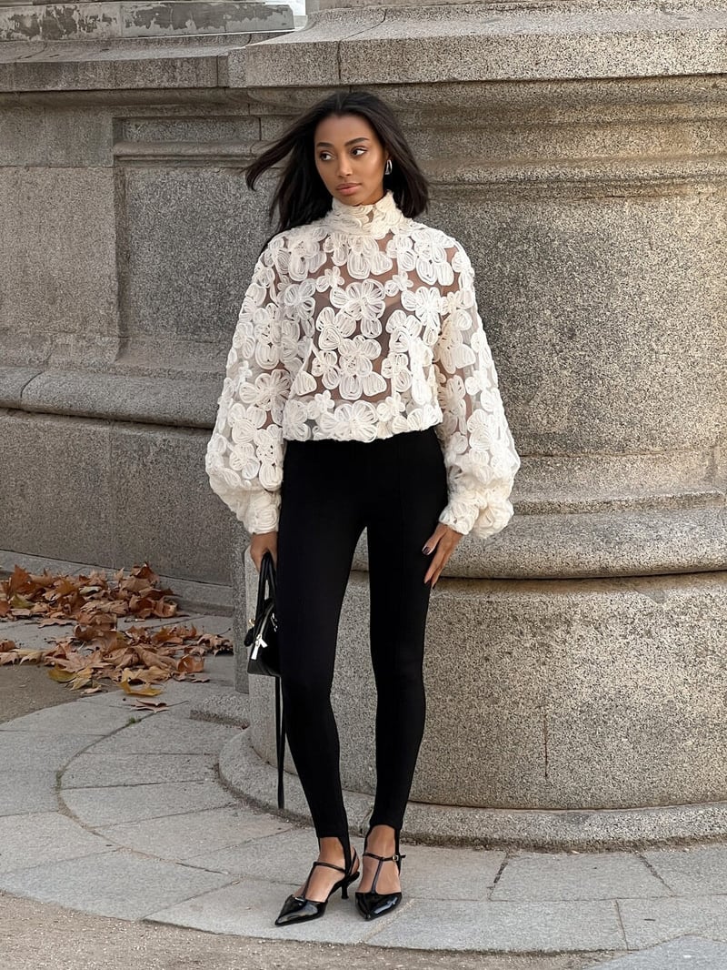 Frau in eleganter, weißer Spitzenbluse und schwarzer Hose steht vor steinernem Hintergrund. Herbstblätter am Boden. Mode, Herbst, Eleganz.