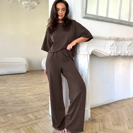 Frau in braunem Lounge-Set lehnt an weißem Kamin in modernem Wohnzimmer. Bequeme Damenmode, elegantes Design, entspannte Atmosphäre.