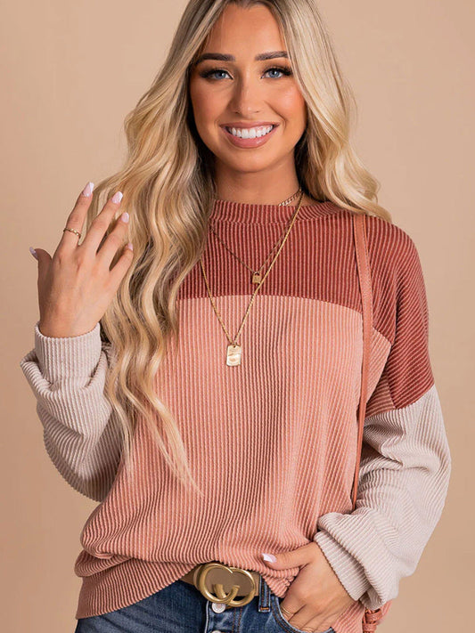 Junge Frau mit langen blonden Haaren trägt gestreiften Pullover in Beige und Rosa, kombiniert mit Jeans und goldenen Halsketten, lächelt in die Kamera.