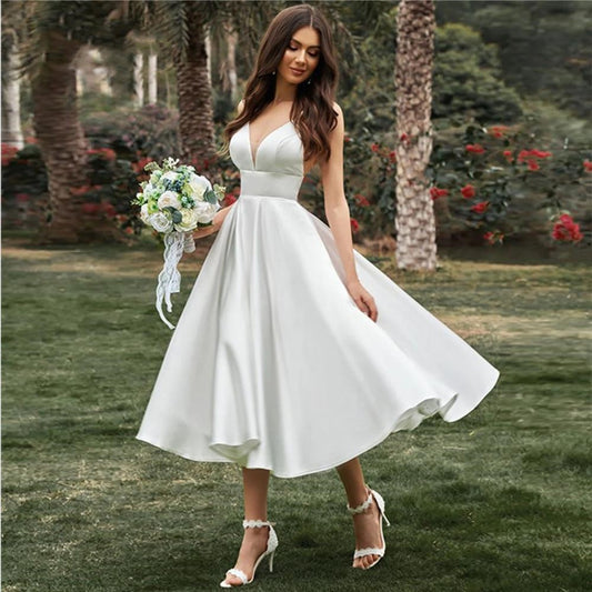 Braut in weißem Hochzeitskleid mit Blumenstrauß im Garten, umgeben von Palmen und Rosen. Elegantes, ärmelloses Design, ideal für Sommerhochzeiten.
