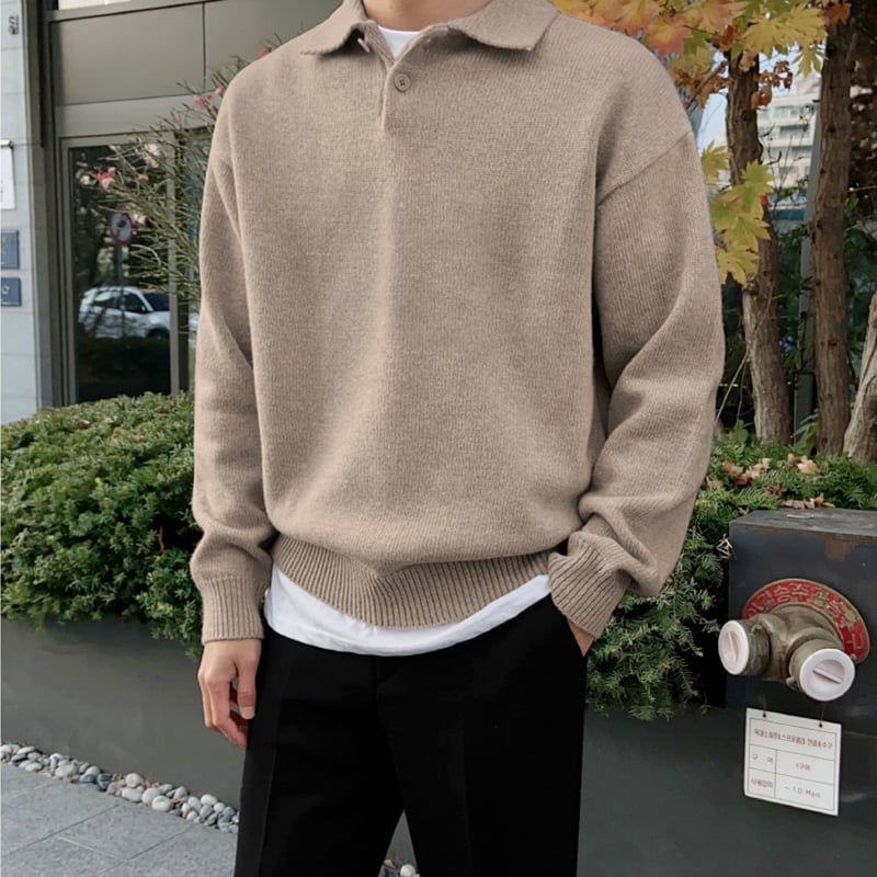 Mann in beigem Pullover und schwarzer Hose steht vor einem Gebäude mit Herbstlaub. Modischer Herbstlook, lässiger Stil, urbane Umgebung.
