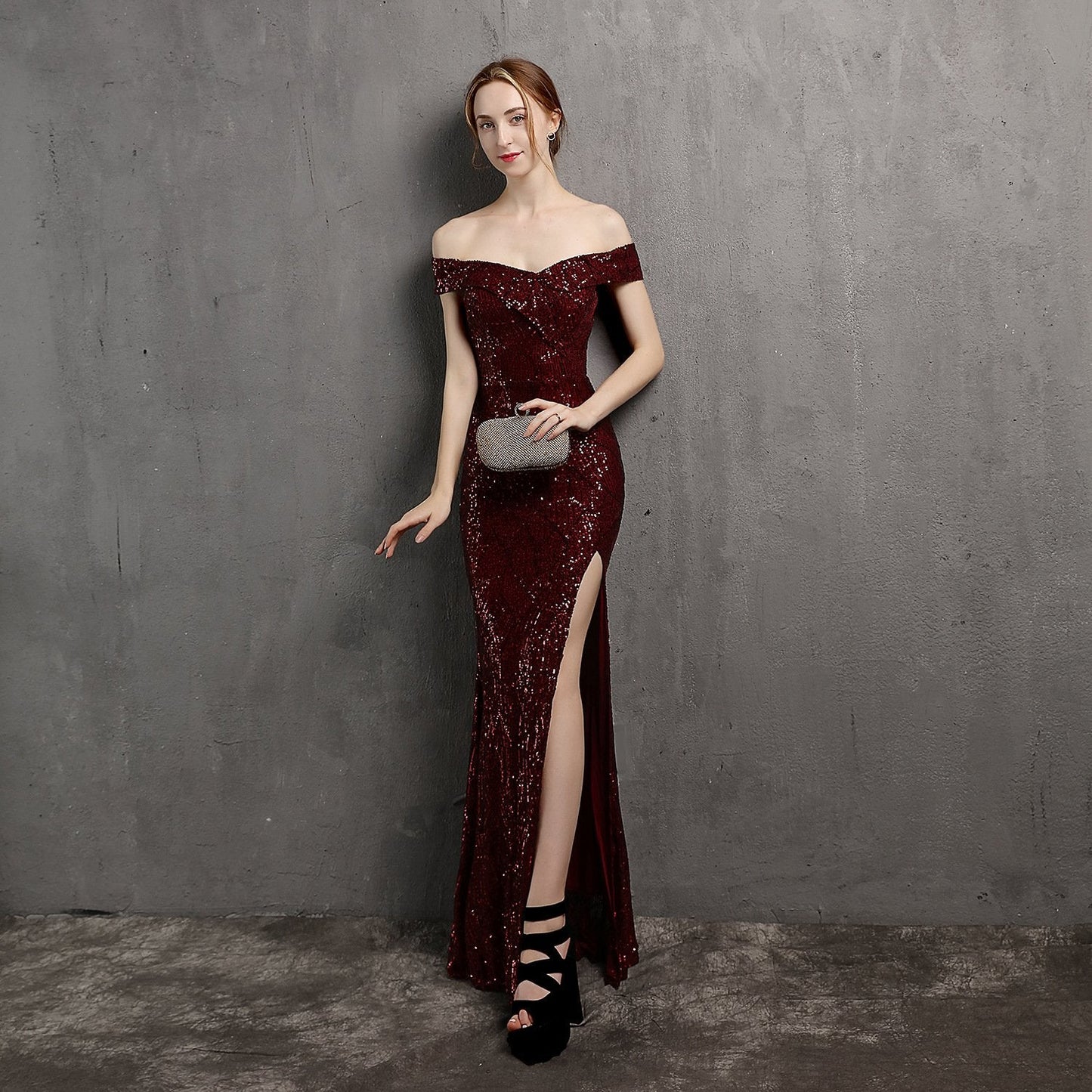 Frau in elegantem, schulterfreiem, weinrotem Paillettenkleid mit hohem Schlitz, hält silberne Clutch, trägt schwarze High Heels, grauer Hintergrund.