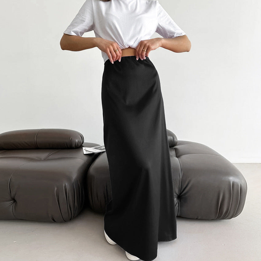 Frau in weißem T-Shirt und schwarzem Maxirock steht vor modernem, grauem Sofa. Minimalistisches Interieur, modisches Outfit, zeitloser Stil.