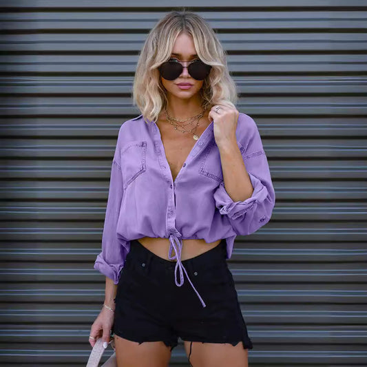 Blonde Frau in lila Bluse und schwarzer Shorts vor grauem Rolltor, trägt Sonnenbrille und Halsketten. Modetrend, Sommeroutfit, Streetstyle.