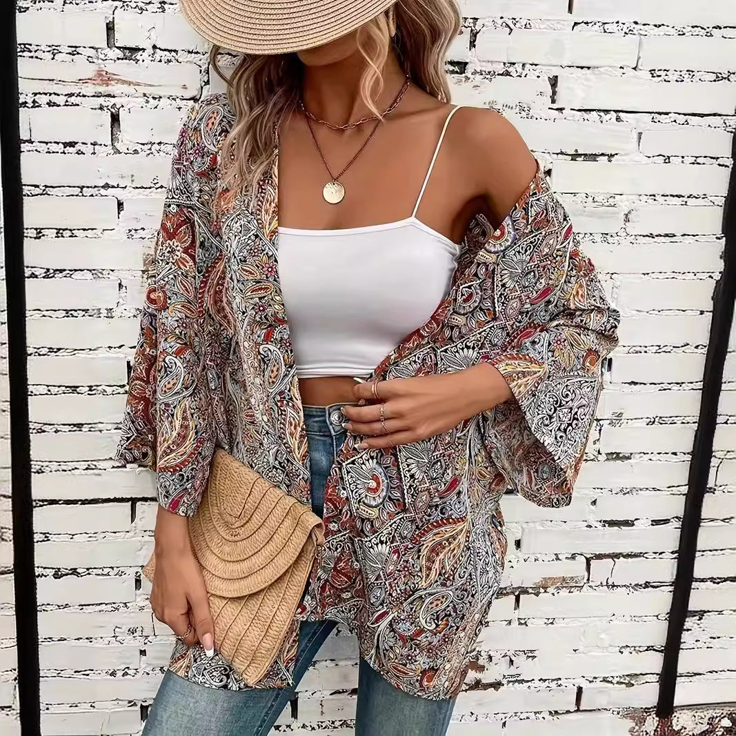 Frau in Boho-Kimono mit Paisley-Muster, weißem Top und Jeans vor weißer Ziegelwand. Strohhut und geflochtene Clutch als Accessoires.