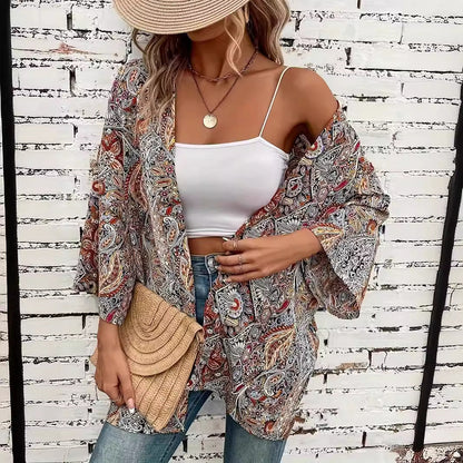 Frau in Boho-Kimono mit Paisley-Muster, weißem Top und Jeans vor weißer Ziegelwand. Strohhut und geflochtene Clutch als Accessoires.