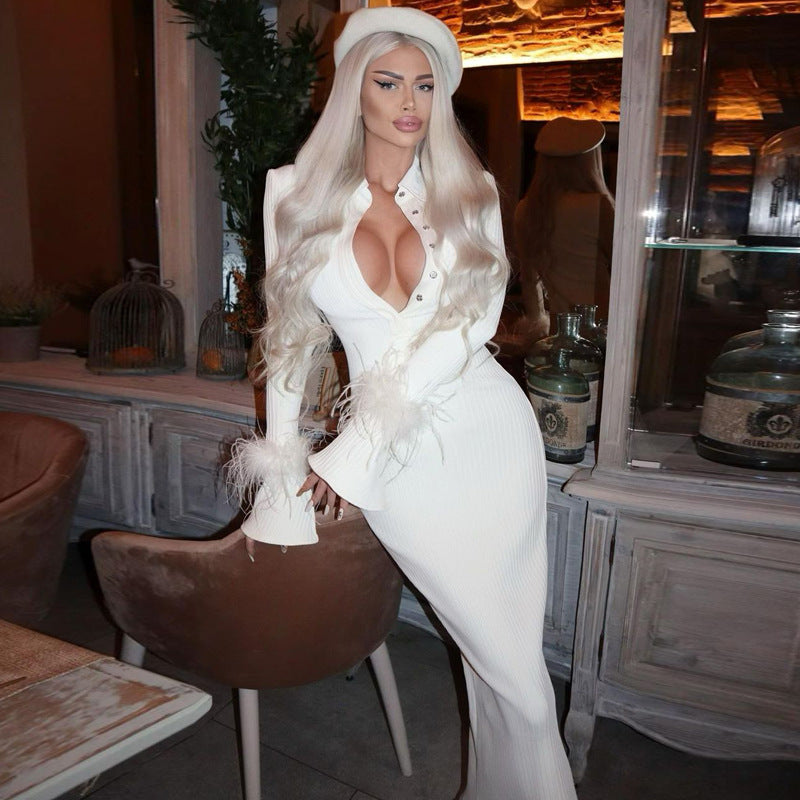 Eine Frau in einem eleganten weißen Kleid mit tiefem Ausschnitt und langen blonden Haaren posiert in einem stilvollen, modern eingerichteten Raum.