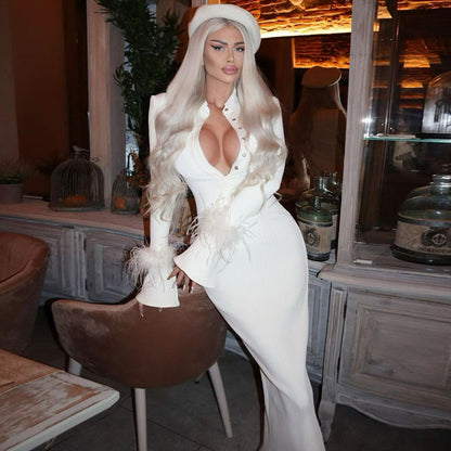 Eine Frau in einem eleganten weißen Kleid mit tiefem Ausschnitt und langen blonden Haaren posiert in einem stilvollen, modern eingerichteten Raum.