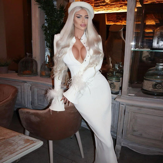 Eine Frau in einem eleganten weißen Kleid mit tiefem Ausschnitt und langen blonden Haaren posiert in einem stilvollen, modern eingerichteten Raum.