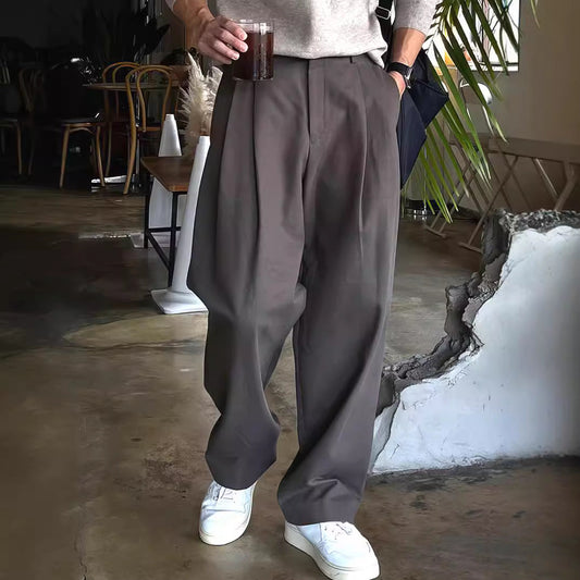 Mann in grauer, weiter Hose und weißen Sneakern hält ein Getränk. Moderne Herrenmode, lässiger Stil, Café-Hintergrund.