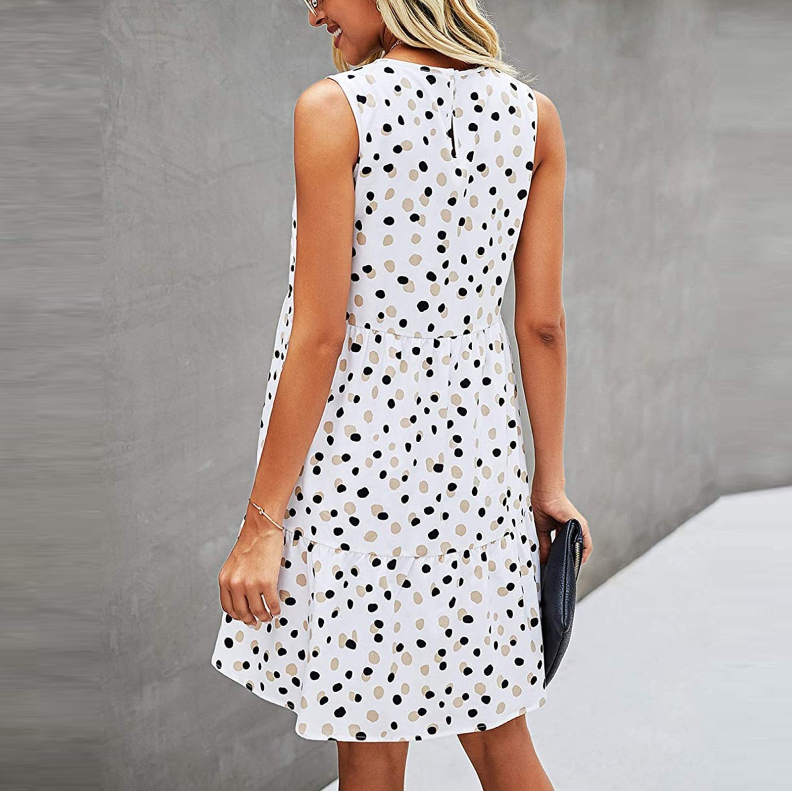 Frau in ärmellosem, weißem Kleid mit schwarzen Punkten, seitlich stehend, grauer Hintergrund. Sommermode, elegantes Design, Freizeitkleidung.