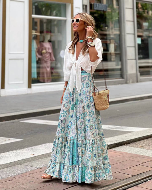 Frau in weißer Bluse und langem, geblümtem Rock auf Stadtstraße, trägt Sonnenbrille und Strohtasche. Sommermode, Streetstyle, Boho-Chic.