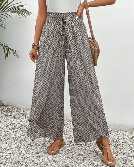 Frau in gemusterten, weiten Sommerhosen, Sandalen und weißem Top, steht auf Kiesweg. Modetrend, Damenmode, lässiger Sommerlook.