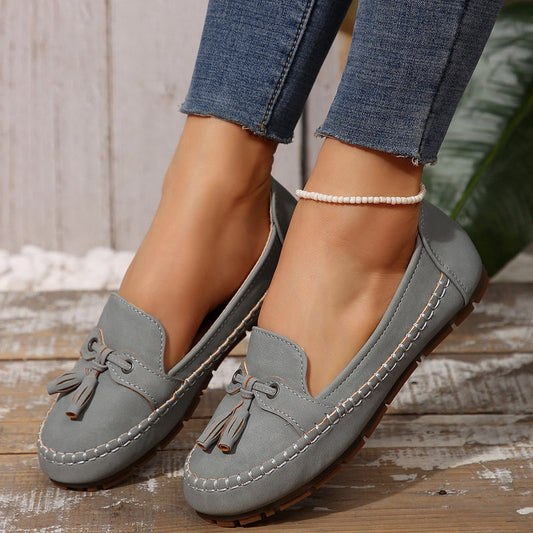 Graue Mokassins mit Quasten, getragen von einer Person in blauen Jeans. Modische Damen-Slipper, bequem und stilvoll, auf Holzboden.