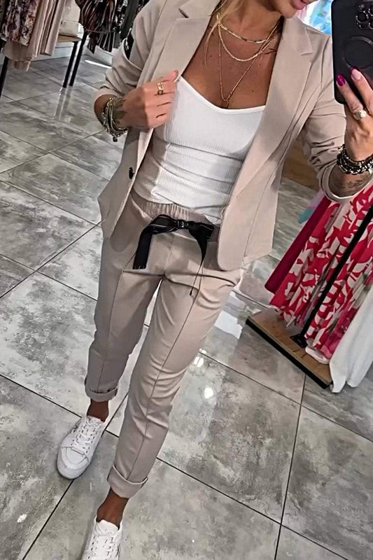 Frau in beigem Anzug mit weißem Top und Sneakers, steht auf glänzendem Fliesenboden in einem Modegeschäft, hält ein Smartphone.