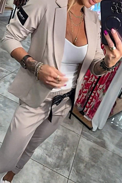Frau in beigem Anzug mit weißem Top, hält Smartphone. Modischer Business-Look, elegante Kleidung, moderne Accessoires, Spiegel-Selfie.