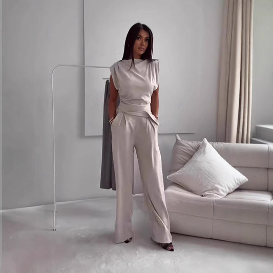 Frau in eleganter, cremefarbener Hose und Oberteil steht in modernem Wohnzimmer mit weißem Sofa und Vorhängen. Minimalistisches Interieur.