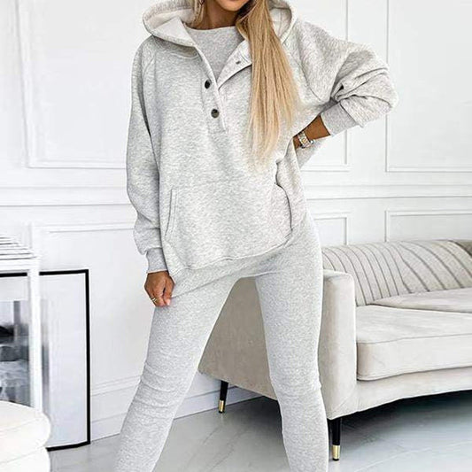 Graues Damen-Sweatshirt mit Kapuze und Knöpfen, kombiniert mit passender Jogginghose. Bequeme Freizeitmode im modernen Wohnzimmer.