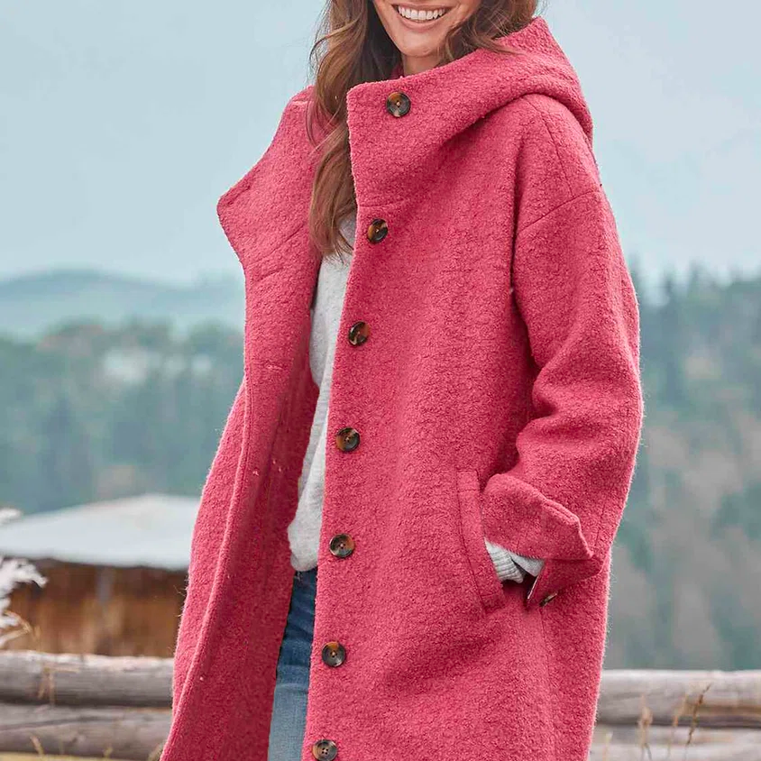 Frau in rosa Wollmantel mit Kapuze, lächelt in ländlicher Umgebung. Modetrend Herbst/Winter, stilvoller Damenmantel, Outdoor-Bekleidung.