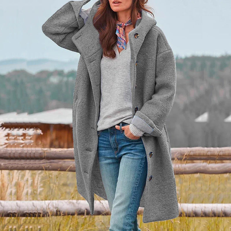 Frau in grauem Wollmantel, Jeans und Schal steht vor ländlicher Kulisse. Herbstmode, Damenbekleidung, Outdoor-Stil.