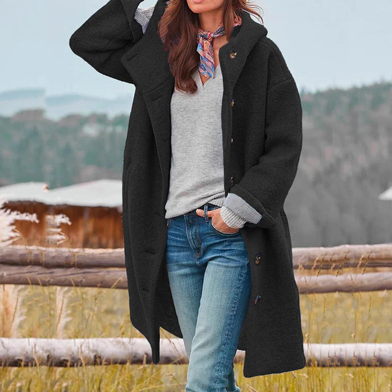 Frau in schwarzem Wollmantel, grauem Pullover und Jeans vor ländlicher Kulisse. Herbstmode, stilvoll, lässig, Outdoor-Bekleidung.