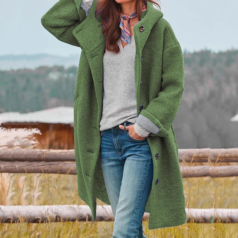 Frau in grünem Wollmantel, grauem Pullover und blauer Jeans vor ländlicher Kulisse. Herbstmode, Outdoor-Bekleidung, stilvoller Look.