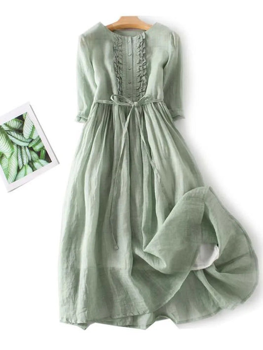 Grünes Leinenkleid mit Rüschen und Bindegürtel, langärmelig, fließend, auf Holzbügel, neben Pflanzenbild, elegantes Sommerkleid.