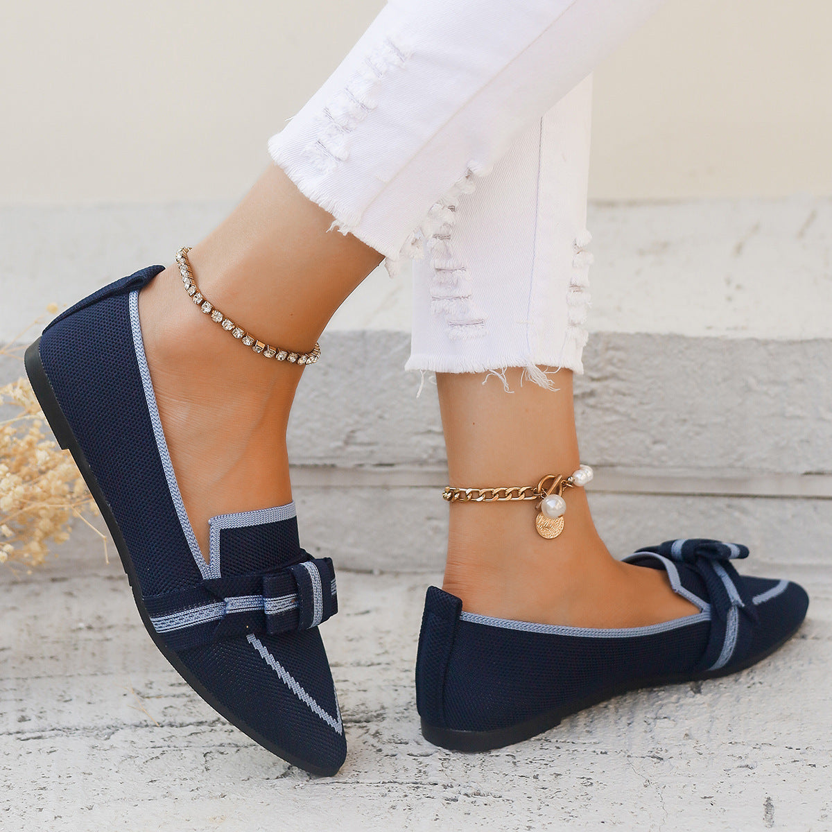 Damenfüße in weißen Jeans und dunkelblauen Loafers mit Schleife, Schmuck an den Knöcheln, auf hellem Hintergrund. Modische Schuhe, Sommerstil.