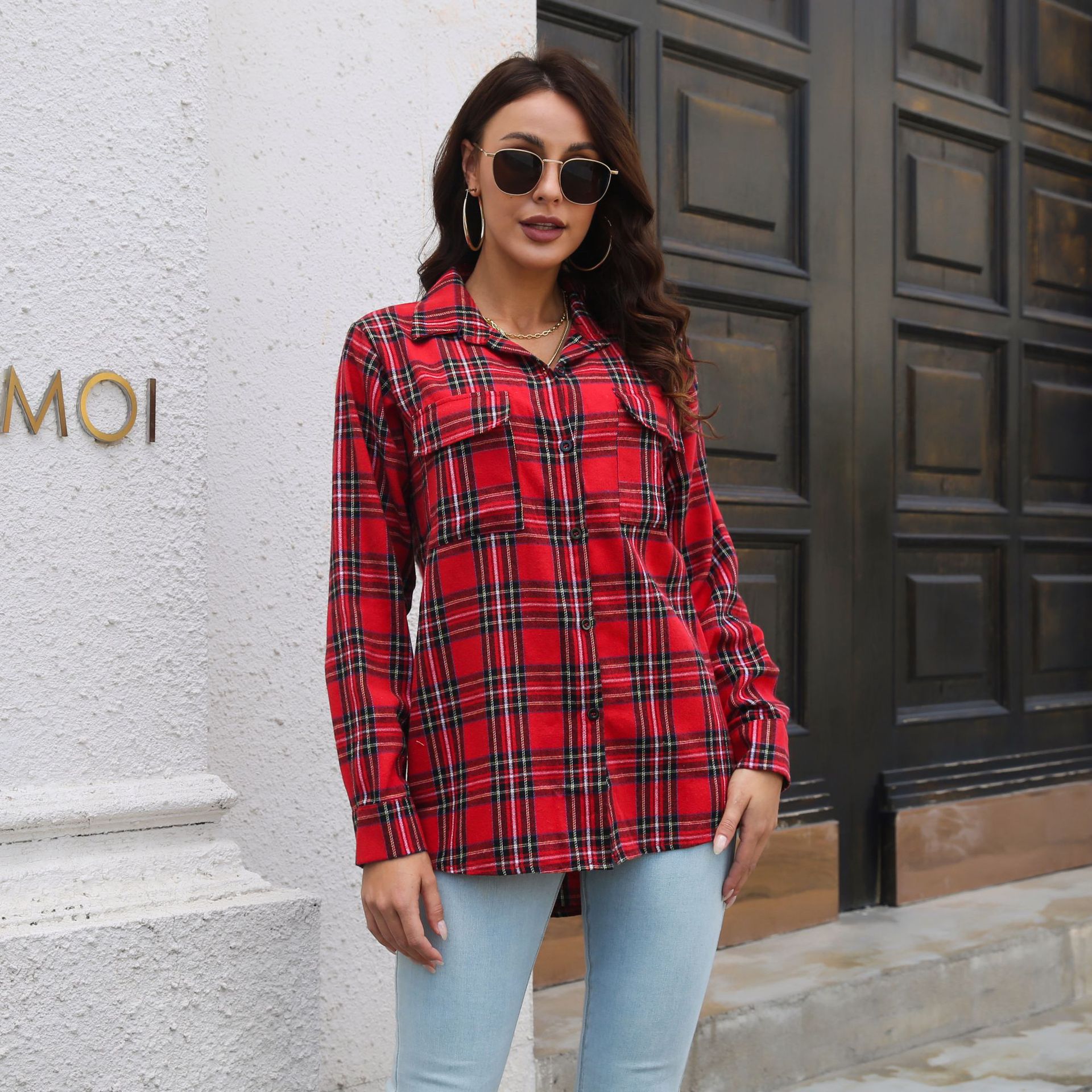 Frau in rotem Karohemd und blauer Jeans vor Holztür, trägt Sonnenbrille und Ohrringe. Modetrend, lässiger Stil, Herbstmode.