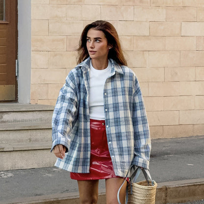 Frau in kariertem Hemd und rotem Lederrock, trägt Strohtasche. Urbaner Hintergrund, modischer Streetstyle-Look.