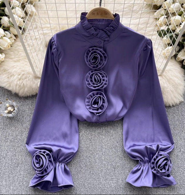 Lila Bluse mit langen Ärmeln, Rüschen und Blumenverzierungen, elegant und modisch, auf grauem Hintergrund mit weißen Blumen im Hintergrund.