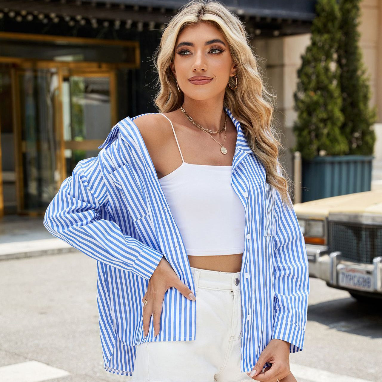 Junge Frau mit blonder Frisur trägt blau-weiß gestreiftes Hemd und weißes Top, steht vor einem Gebäude. Modetrend, Streetstyle, Sommermode.